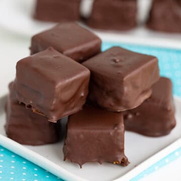 A stack of chocolate-covered cheesecake bites is artfully arranged on a white plate, with more treats in the background, resting elegantly on a turquoise napkin.