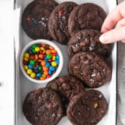 A hand reaches for double chocolate cookies studded with vibrant M&M pieces, arranged in a white tray beside a small bowl of candy-coated chocolates.