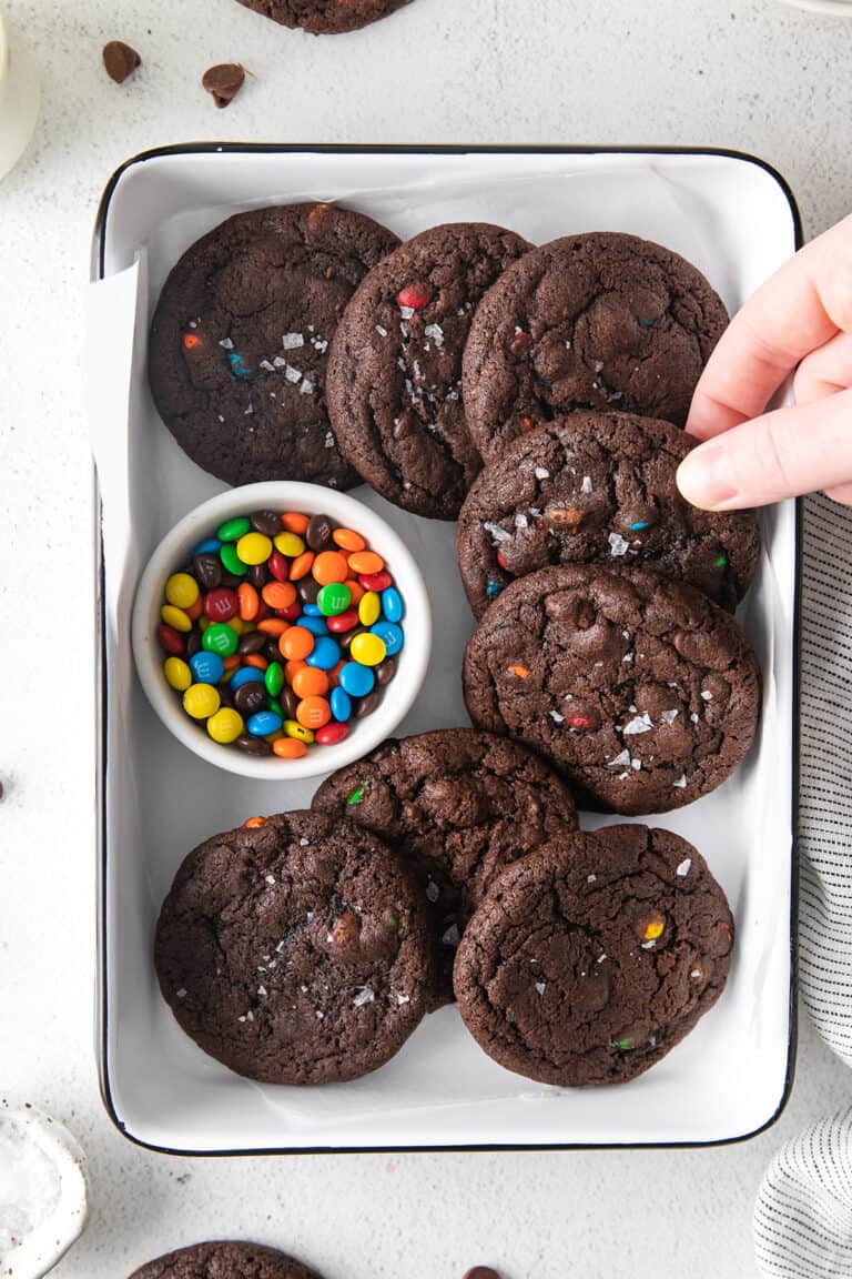 A hand reaches for double chocolate cookies studded with vibrant M&M pieces, arranged in a white tray beside a small bowl of candy-coated chocolates.