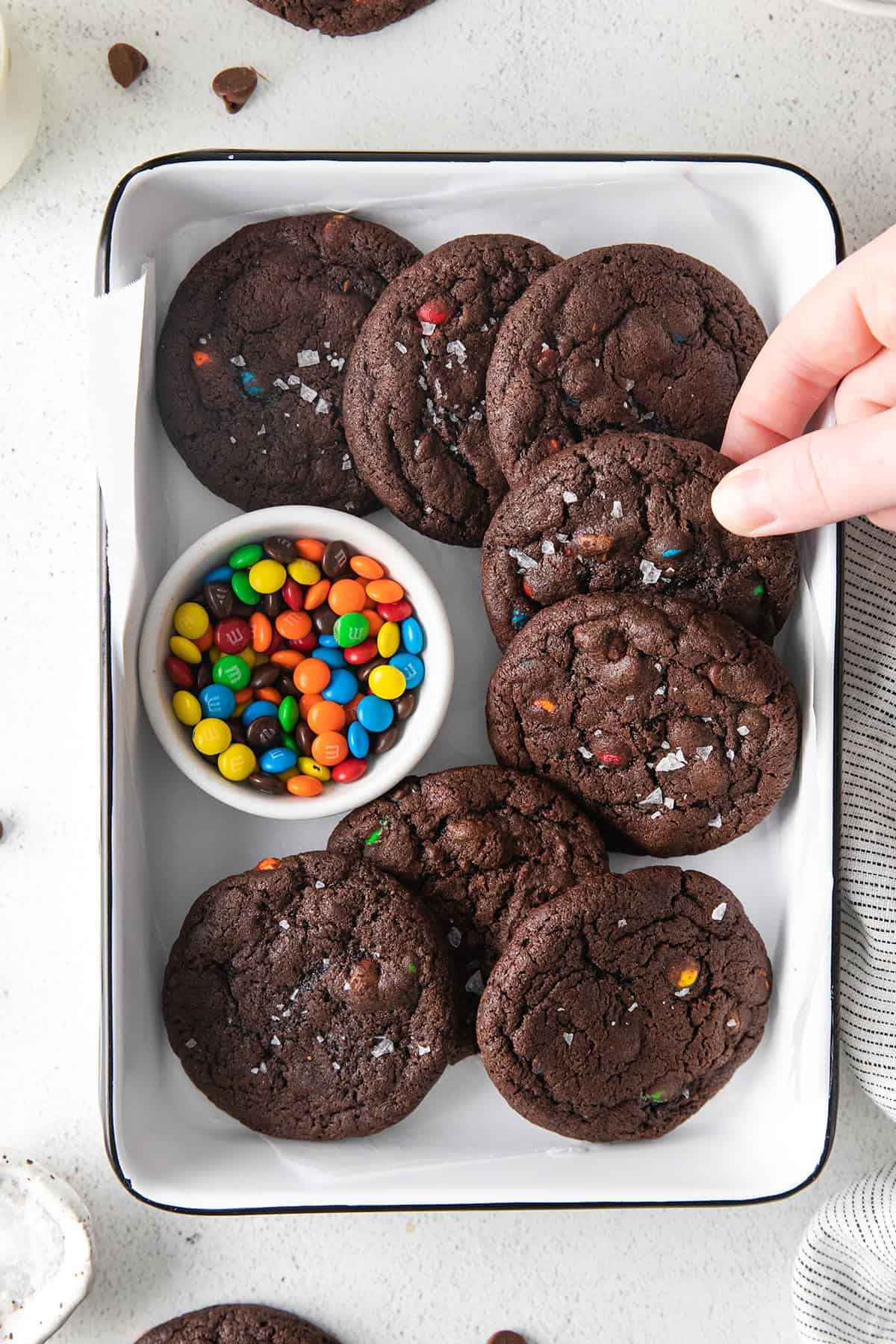 A hand reaches for double chocolate cookies studded with vibrant M&M pieces, arranged in a white tray beside a small bowl of candy-coated chocolates.