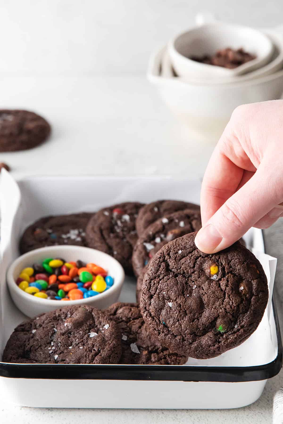 A hand holds a chocolate cookie studded with vibrant M&M candies, hovering above a tray of similar cookies and a bowl brimming with more colorful treats.