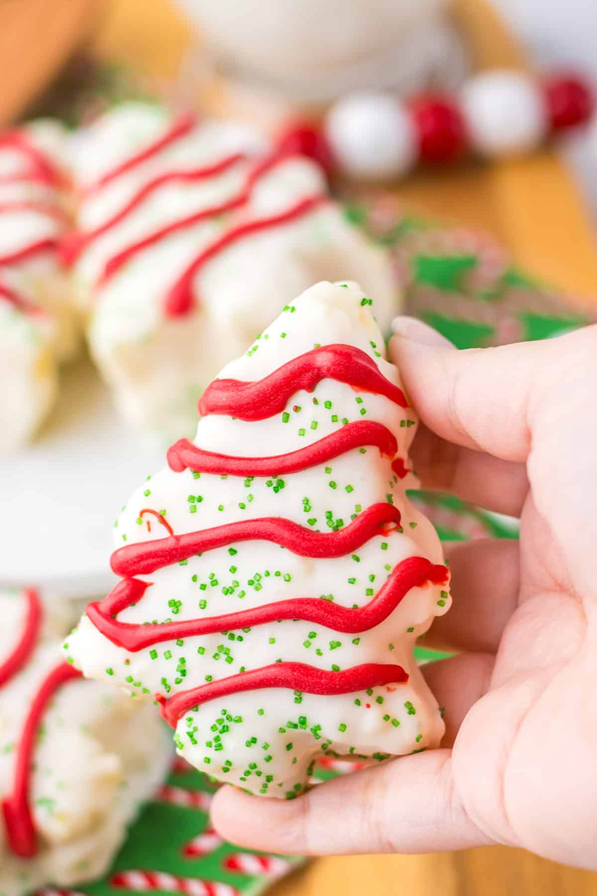 A hand holding a delightful Christmas tree-shaped cookie, reminiscent of Little Debbie Christmas Tree Cakes, adorned with white icing, red frosting stripes, and a sprinkle of green.