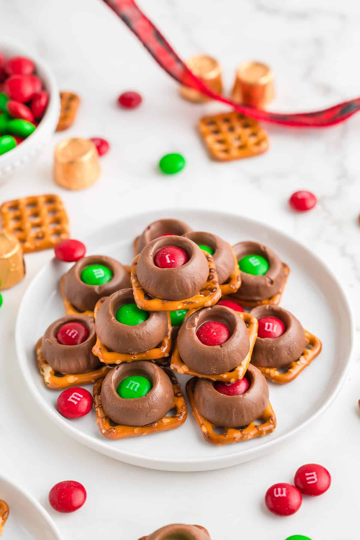 A plate of delightful pretzel treats topped with chocolate, caramel, and vibrant red and green candy-coated chocolates, featuring a hidden Rolo surprise, all surrounded by scattered ingredients on a luxurious marble surface.