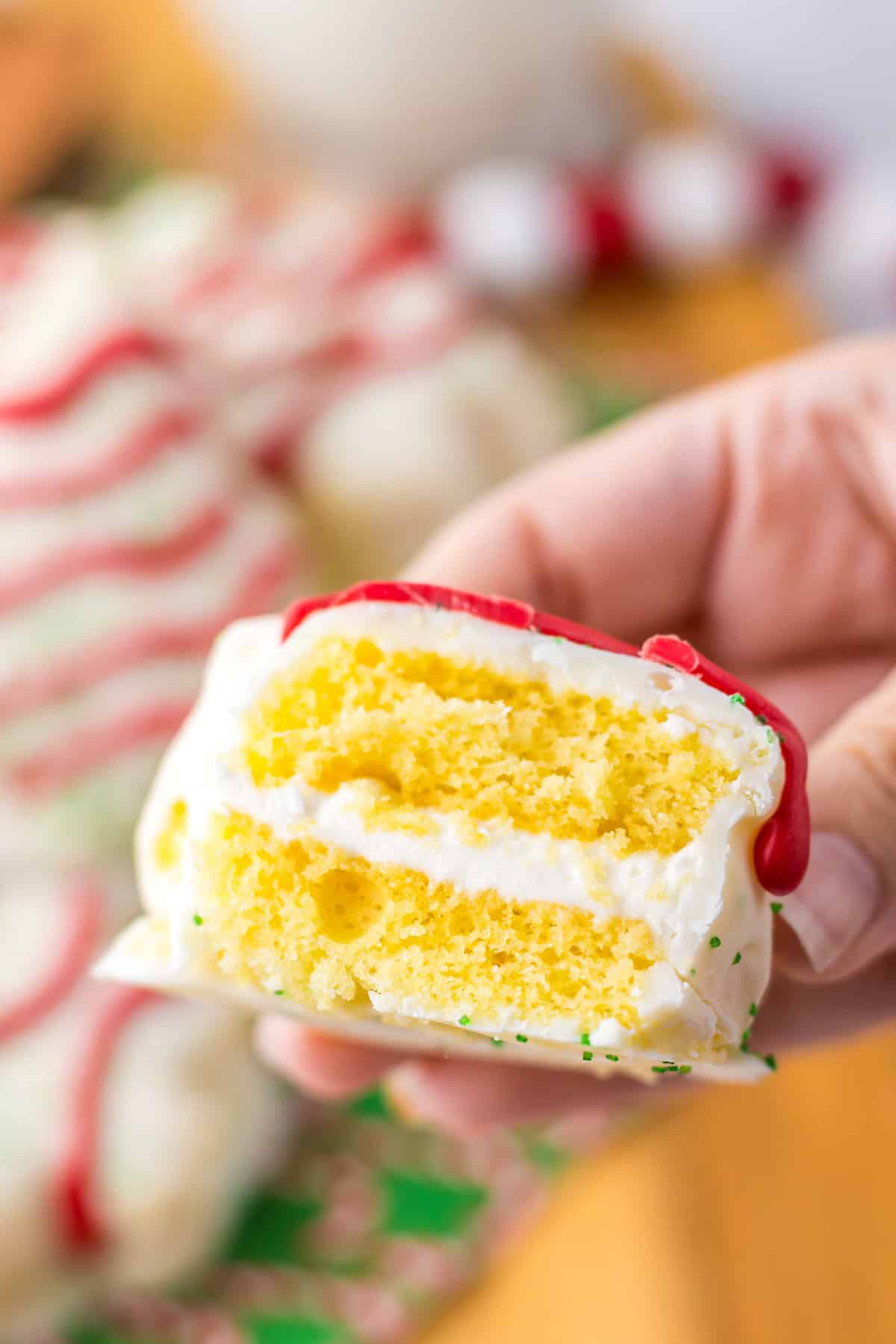 A hand holds a slice of yellow cake with white frosting, reminiscent of Little Debbie Christmas Tree Cakes, elegantly decorated with red icing and green sprinkles.