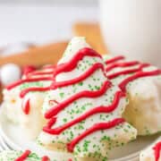 A festive plate of Christmas tree-shaped cookies with red icing and green sprinkles, resembling Little Debbie's iconic treats, sits alongside a glass of milk in the background.