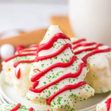 A festive plate of Christmas tree-shaped cookies with red icing and green sprinkles, resembling Little Debbie's iconic treats, sits alongside a glass of milk in the background.