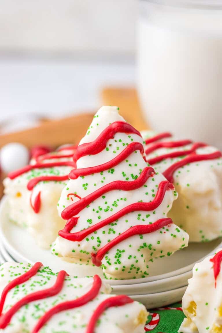 A festive plate of Christmas tree-shaped cookies with red icing and green sprinkles, resembling Little Debbie's iconic treats, sits alongside a glass of milk in the background.