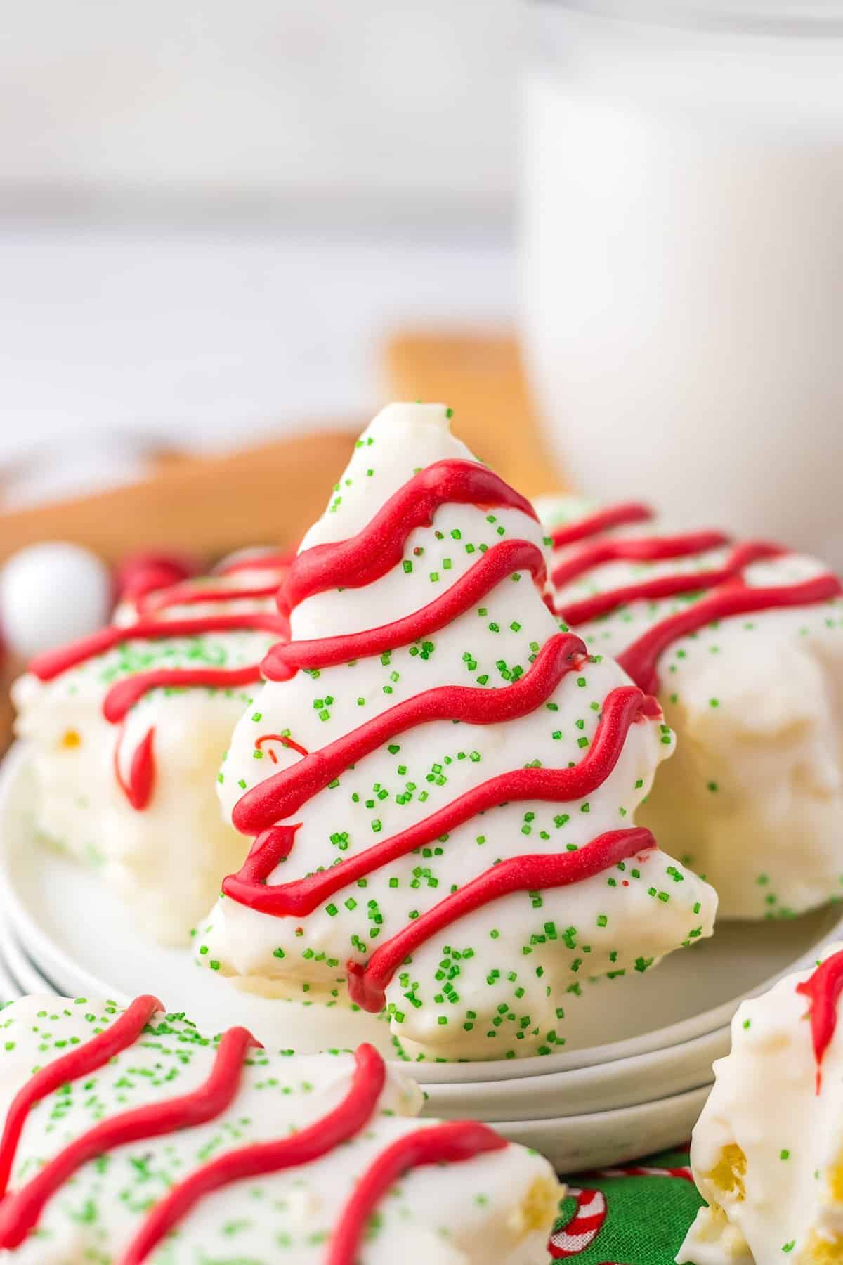 A festive plate of Christmas tree-shaped cookies with red icing and green sprinkles, resembling Little Debbie's iconic treats, sits alongside a glass of milk in the background.