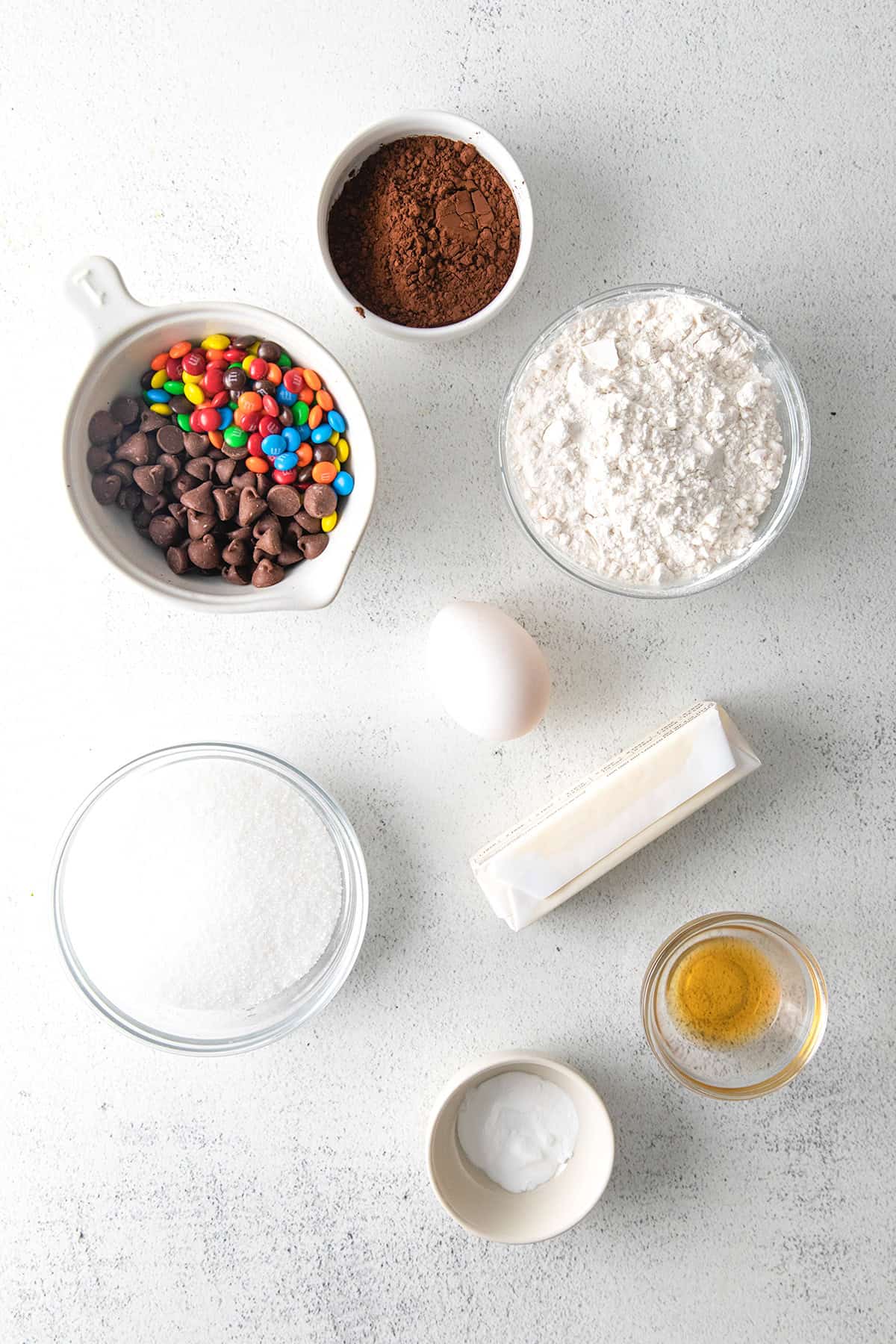 Assorted baking ingredients including chocolate candies, cocoa powder, flour, sugar, egg, butter, baking soda, and vanilla extract on a white surface—perfect for whipping up delicious cookies.