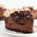 A slice of chocolate cake with decadent layers resembling a chocolate Oreo cheesecake, topped with Oreo cookie crumbles and luscious cream, elegantly presented on a white plate.