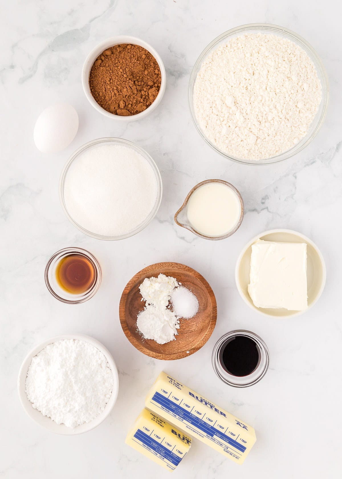 A collection of baking ingredients for delightful Red Velvet treats on a marble surface, including flour, cocoa, sugar, vanilla, an egg, milk, butter, cream cheese, baking soda, and salt.