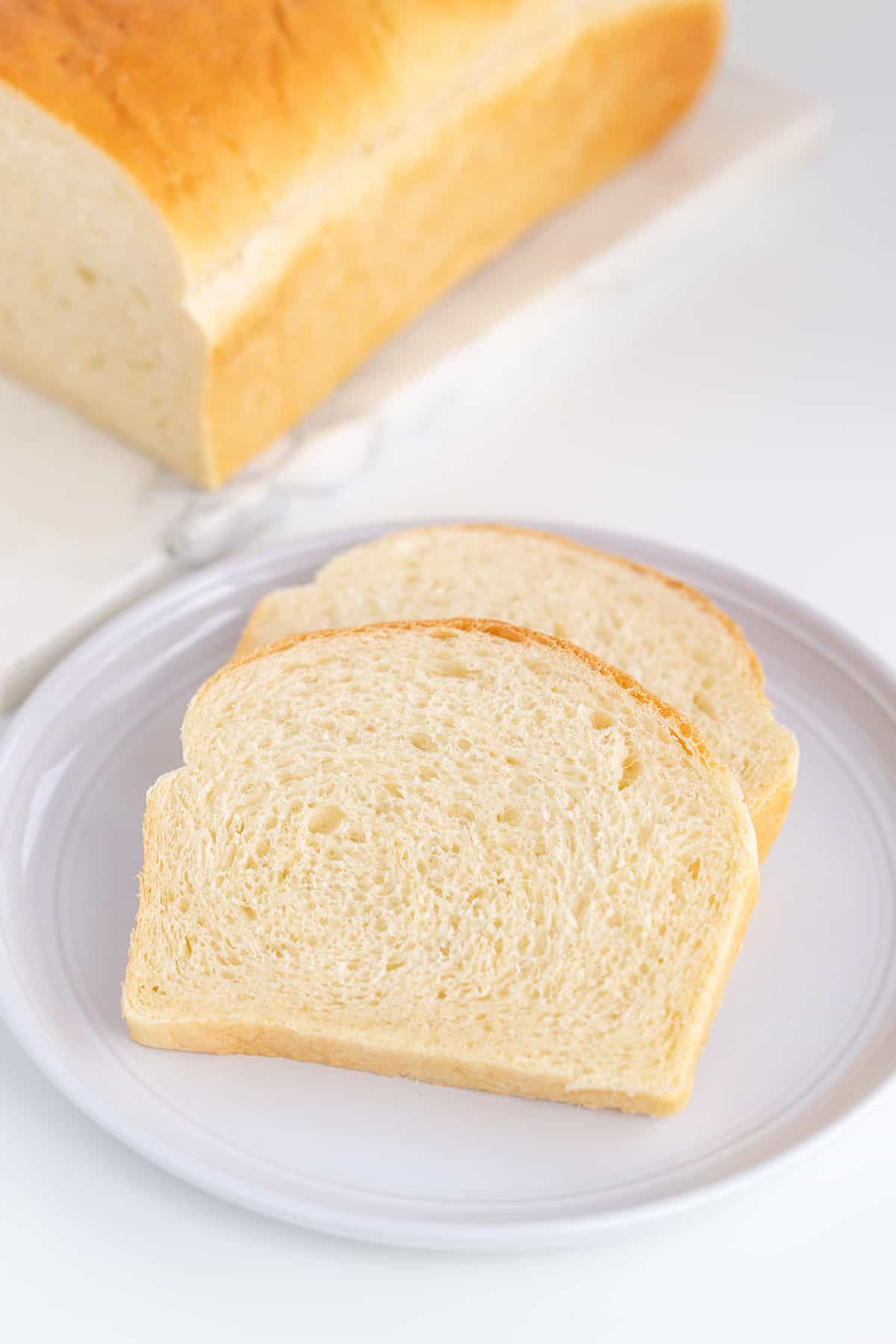 Two slices of white potato bread rest on a white plate, with a loaf in the background.