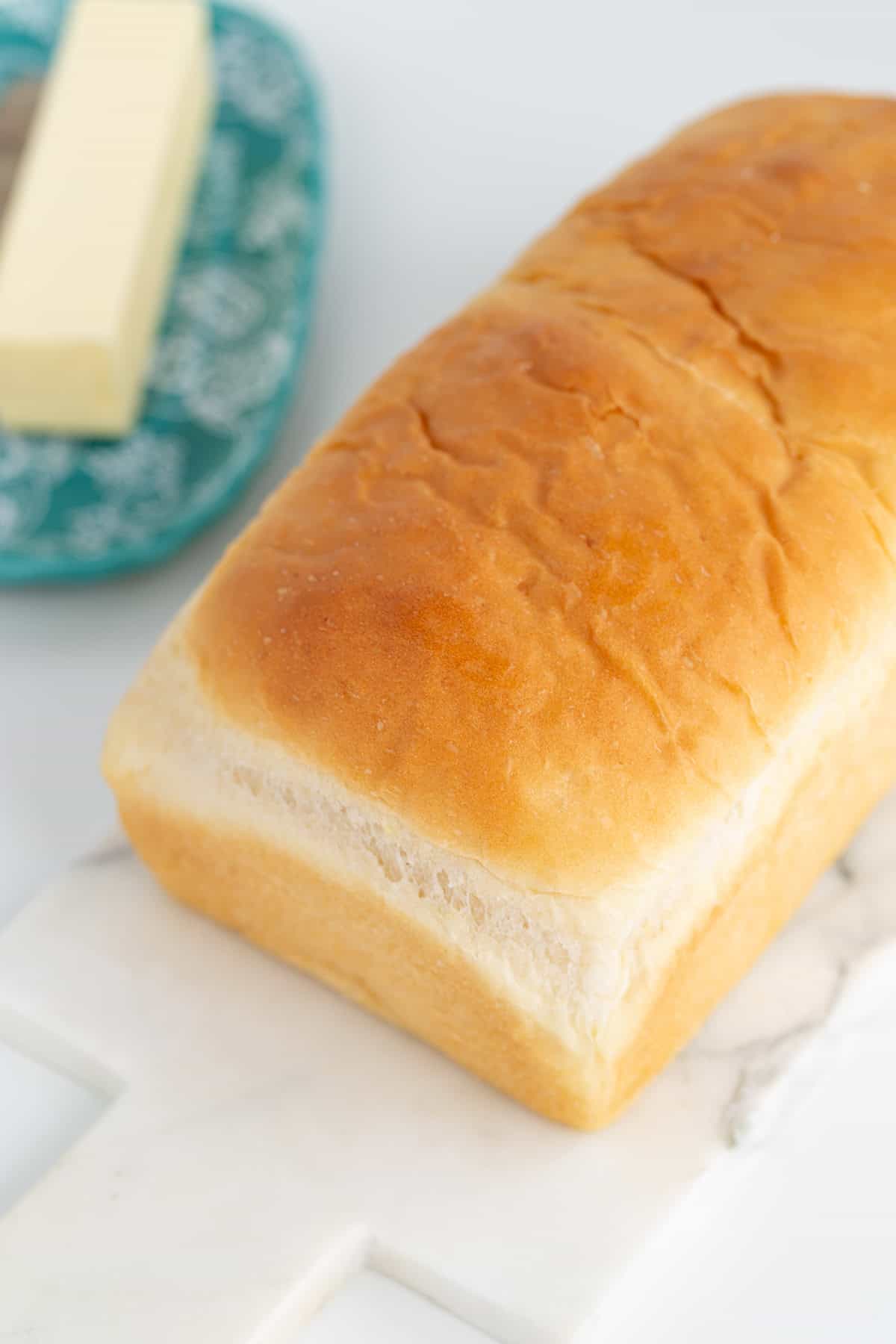 A loaf of potato bread sits on a marble cutting board, with a stick of butter elegantly placed on a blue patterned plate in the background.
