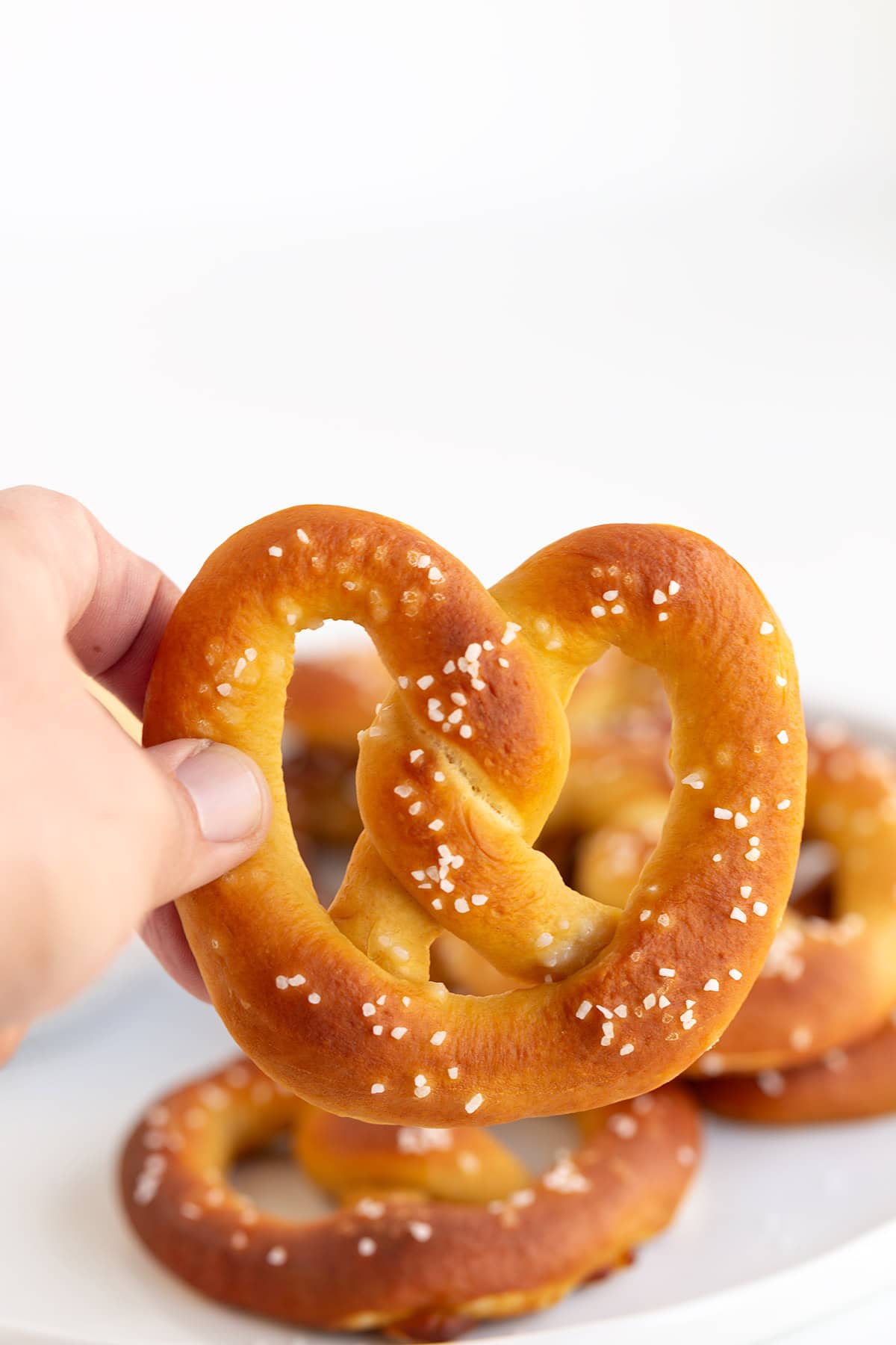 A hand holding a homemade, salted pretzel with more soft pretzels in the background.