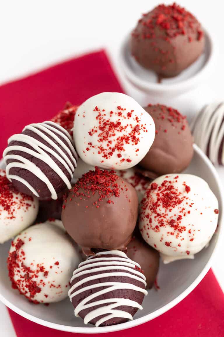 A plate of red velvet cake pops, some coated in white chocolate with red sprinkles, others in milk chocolate, placed on a red napkin. Each bite reveals the smooth taste of cream cheese.