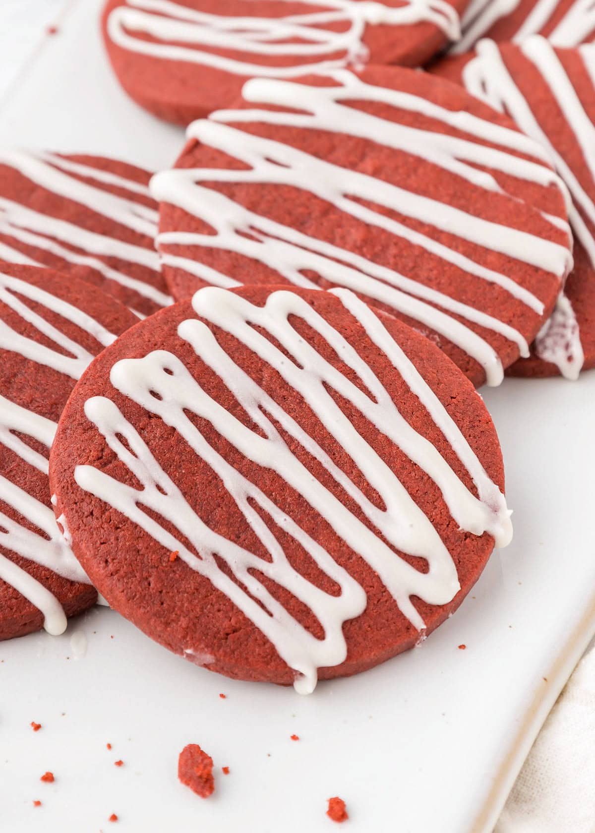 Red velvet cookies with white icing drizzles rest delicately on a pristine white plate, offering a delightful twist on the traditional sugar cookie.