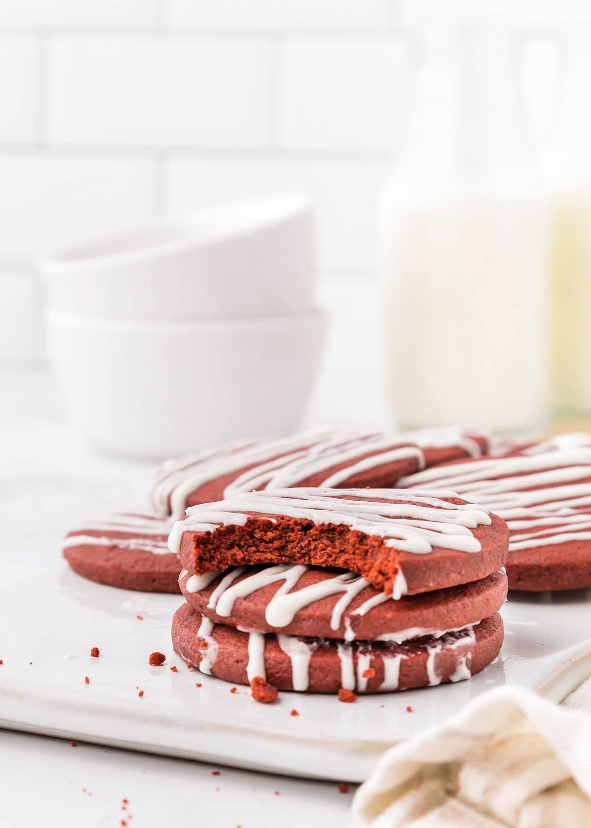 A stack of red velvet cookies with white icing on a white plate tempts the taste buds. One sugar cookie has a bite taken out, hinting at its softness. In the background, bowls and bottles are artfully blurred, adding to the scene's allure.