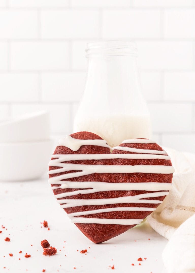 A heart-shaped red velvet cookie with white icing sits in front of a glass bottle of milk on a white surface against a white brick background, offering a delightful twist on classic sugar cookies.
