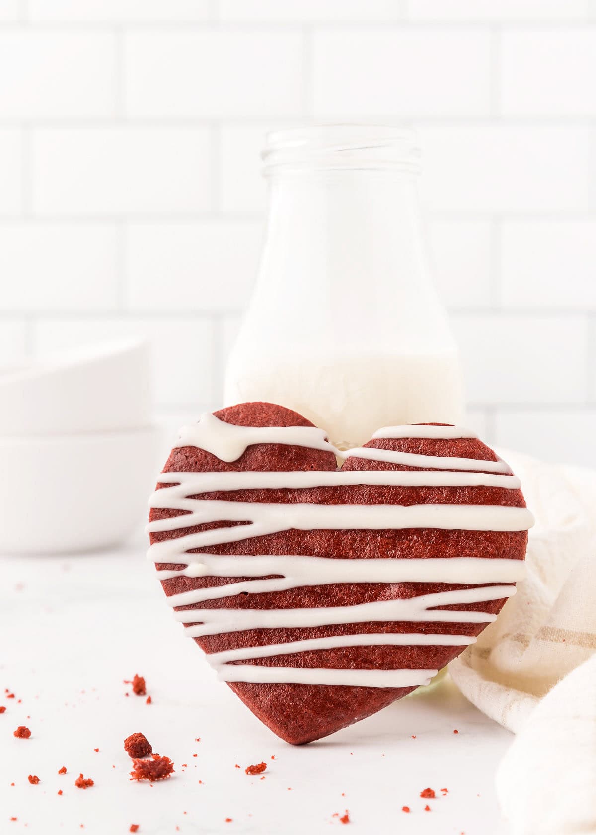 A heart-shaped red velvet cookie with white icing sits in front of a glass bottle of milk on a white surface against a white brick background, offering a delightful twist on classic sugar cookies.