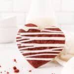A heart-shaped red velvet cookie with a white icing drizzle sits invitingly on a pristine surface, complemented by a glass of milk in the background.