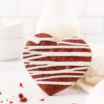 A heart-shaped red velvet cookie with a white icing drizzle sits invitingly on a pristine surface, complemented by a glass of milk in the background.