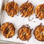 Six homemade Samoa cookies topped with caramel, coconut, and a chocolate drizzle sit on a parchment-lined baking sheet.