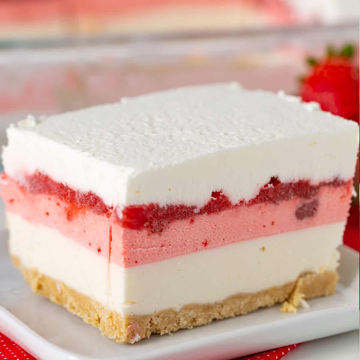 A slice of dessert lasagna with a crumbly base, white and pink creamy layers, and a luscious strawberry topping, served elegantly on a white plate.