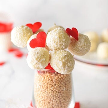 White chocolate coconut truffles on skewers, adorned with red heart cutouts, are artfully arranged in a glass brimming with beige beads. Their delicate presentation on a white surface is complemented by scattered red hearts.