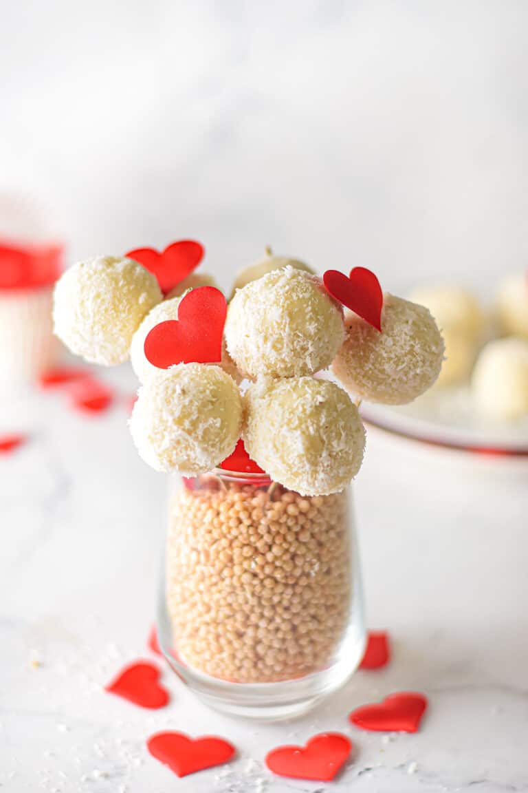 White chocolate coconut truffles on skewers, adorned with red heart cutouts, are artfully arranged in a glass brimming with beige beads. Their delicate presentation on a white surface is complemented by scattered red hearts.