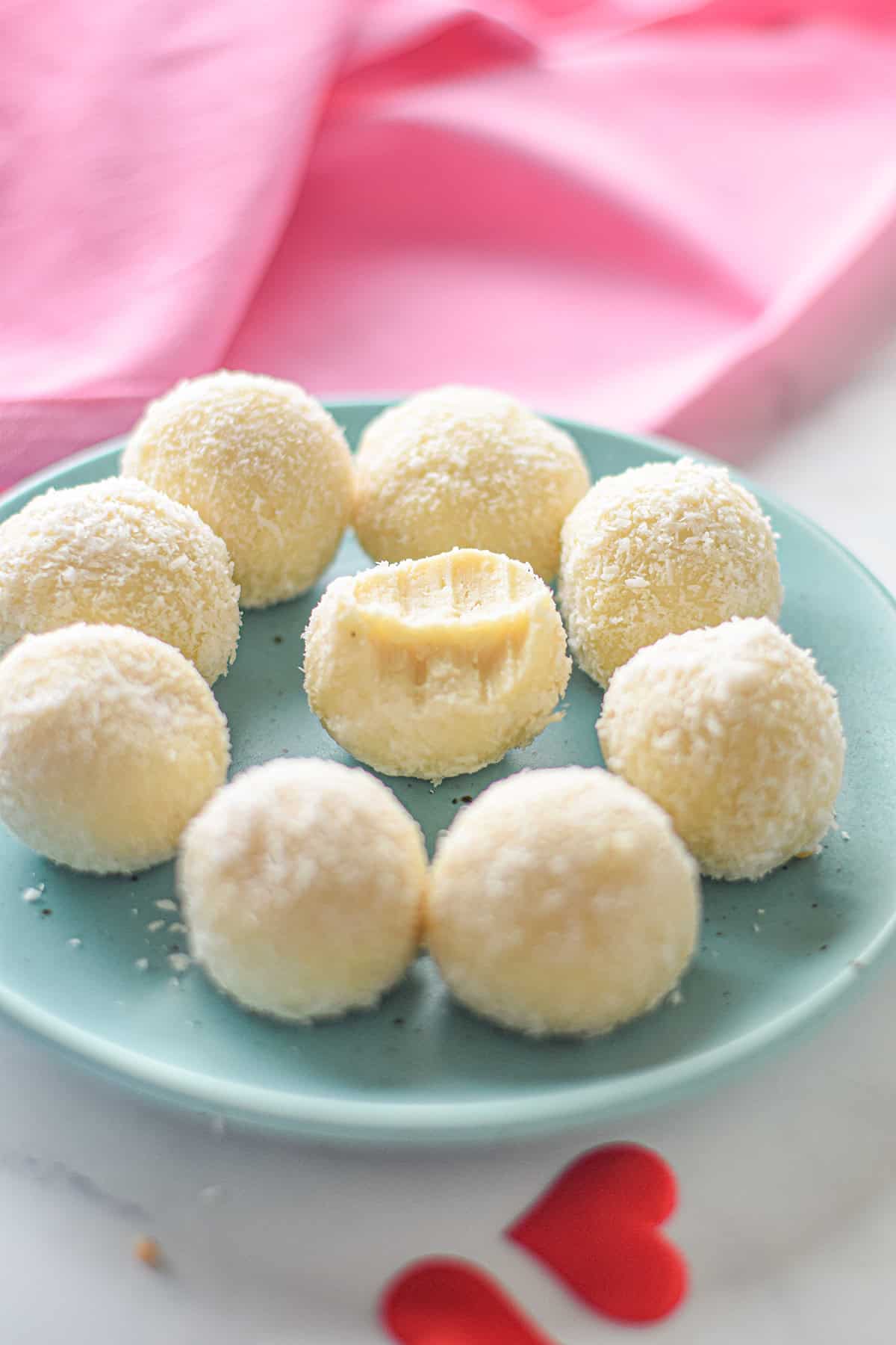 Round coconut-covered truffles on a teal plate, with one revealing a bite. Pink fabric and red heart-shaped cutouts in the background add charm to these white chocolate delights.