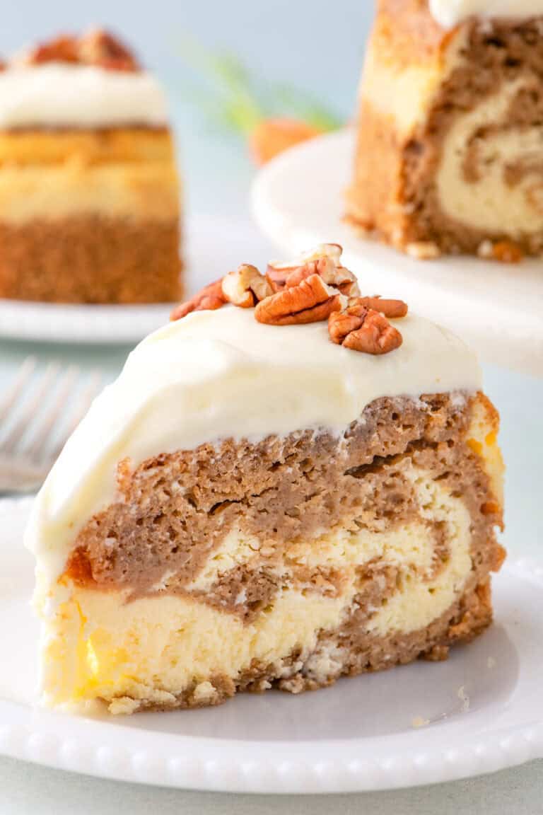 A slice of marble cake with cream cheese frosting and pecans, reminiscent of a luscious cheesecake, sits on a white plate.