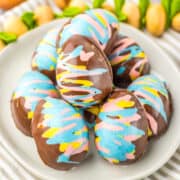 A plate of chocolate-covered marshmallow eggs decorated with blue, pink, and yellow swirls. A wooden spoon and green plant stems are in the background on a striped cloth.