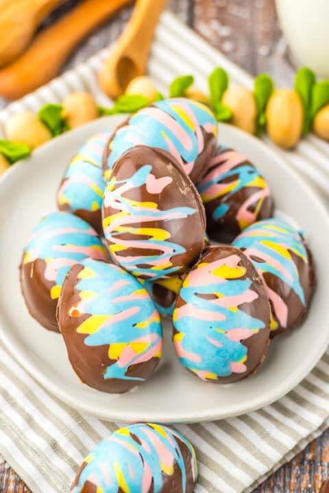 A plate of chocolate-covered marshmallow eggs decorated with blue, pink, and yellow swirls. A wooden spoon and green plant stems are in the background on a striped cloth.