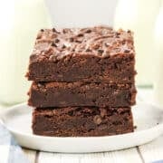 Three stacked eggless brownies on a white plate with bottles of milk in the background, showcasing the delicious art of egg-free baking.