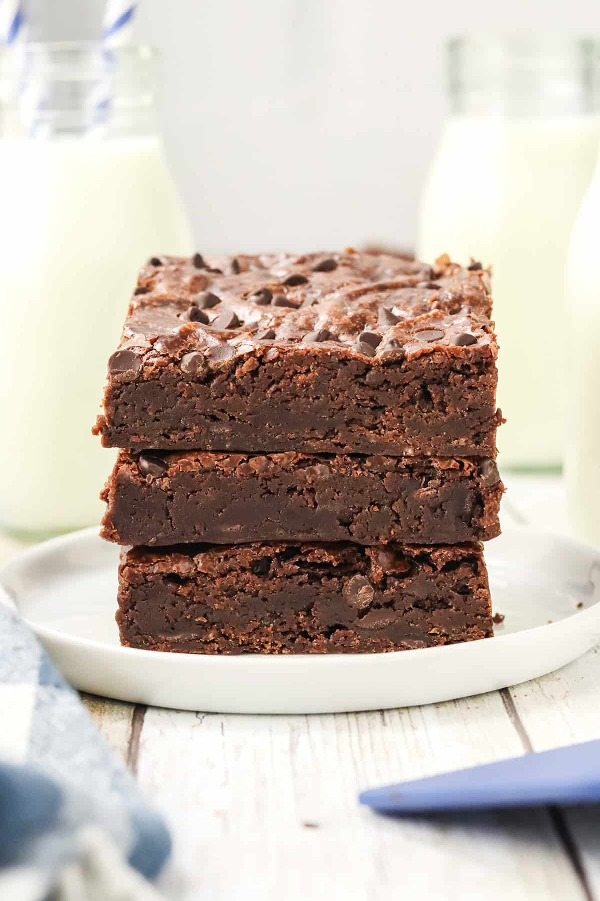 Three stacked eggless brownies on a white plate with bottles of milk in the background, showcasing the delicious art of egg-free baking.