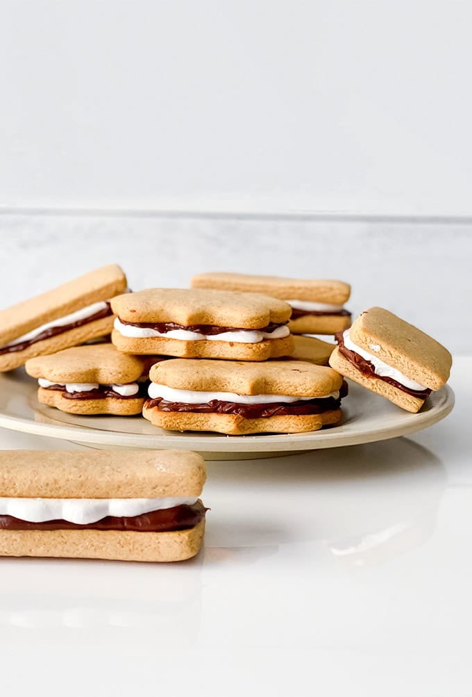 A white plate with multiple stacked sandwich cookies, each filled with chocolate and cream, evokes the classic taste of s'mores as it sits gracefully on a pristine white surface.