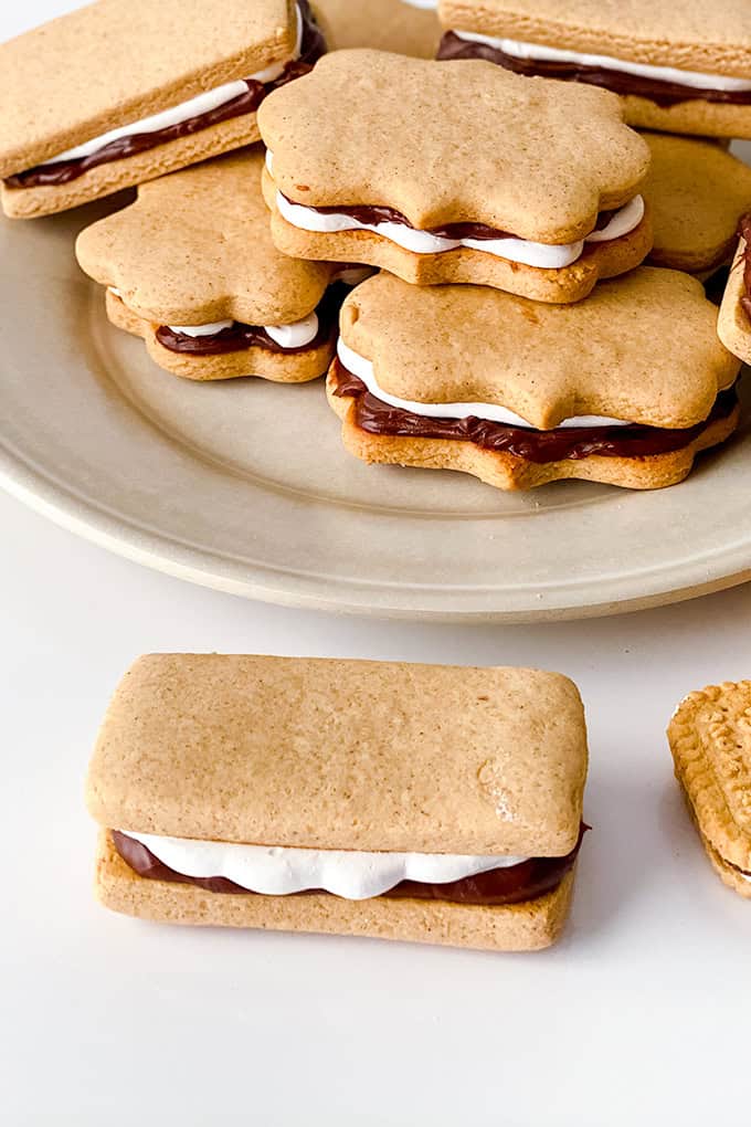 A plate of sandwich-shaped cookies with a rich cream and chocolate filling, reminiscent of classic s'mores, sits invitingly on a white surface, with a rectangular cookie in the foreground.