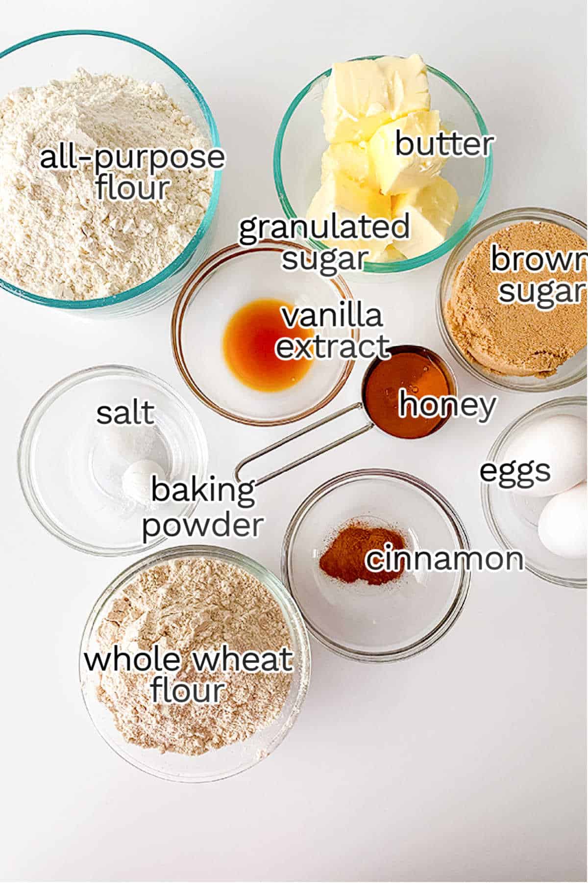 Baking ingredients on a table include all-purpose flour, whole wheat flour, granulated sugar, brown sugar, butter, eggs, vanilla extract, honey, salt, baking powder, and cinnamon—perfect for crafting delicious treats like sandwich cookies or even a twist on classic s'mores.