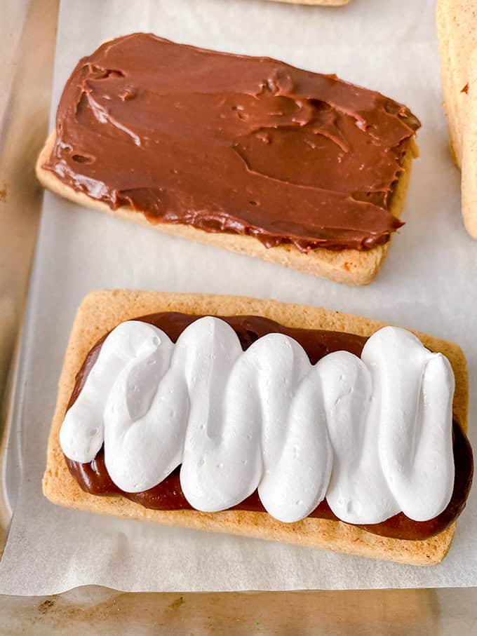Two rectangular pastries rest on parchment paper. One mimics s'mores with a chocolate spread, while the other resembles sandwich cookies, topped with red jam and a white icing zigzag.