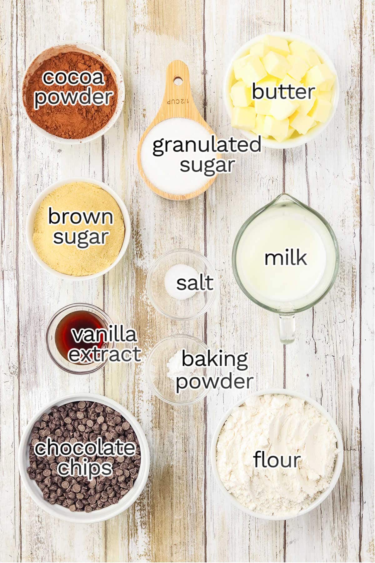 Various baking ingredients arranged on a wooden surface, ready to craft delicious eggless brownies: cocoa powder, butter, granulated sugar, brown sugar, salt, milk, vanilla extract, baking powder, chocolate chips, and flour.