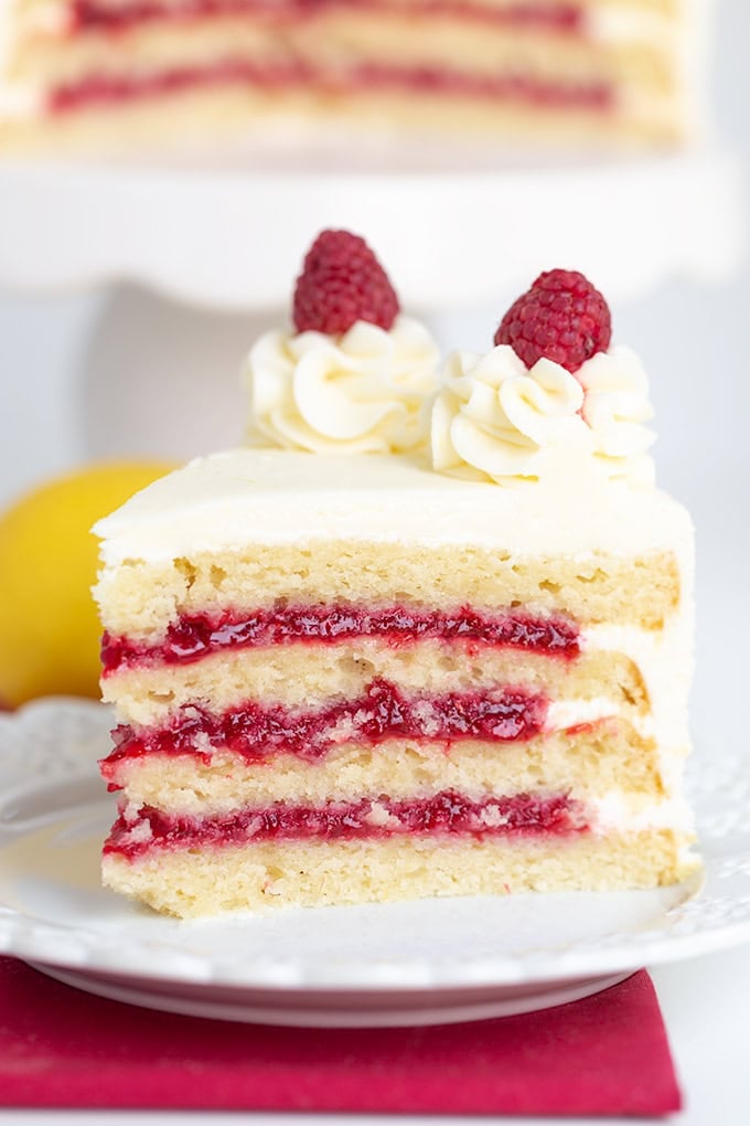 A slice of lemon layer cake with white frosting and raspberry filling, topped with whipped cream and fresh raspberries, rests elegantly on a white plate.