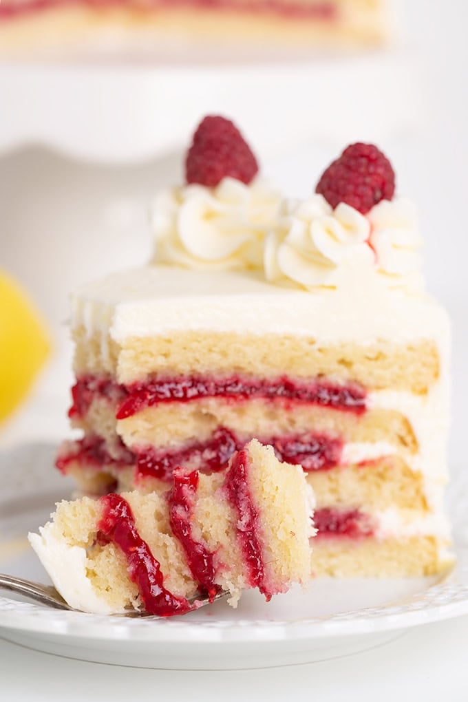 A slice of layered sponge cake with white frosting and raspberry filling, topped with whipped cream and two raspberries. A hint of lemon zest adds a citrus twist. A fork with a piece of cake is on a white plate.