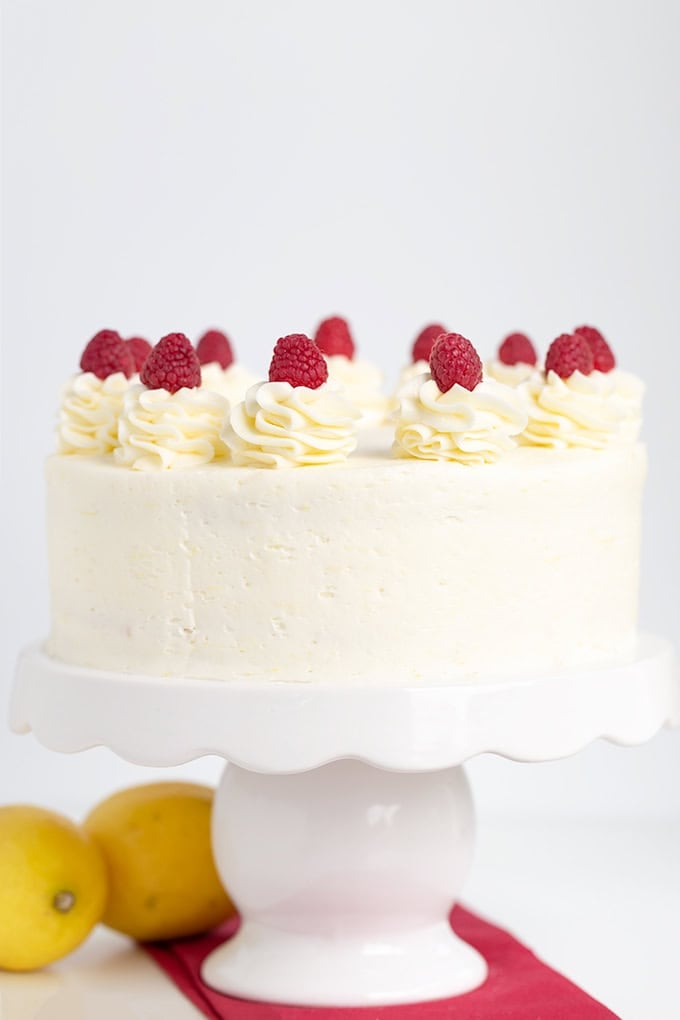 A white frosted cake crowned with raspberry and whipped cream swirls sits elegantly on a white cake stand. Nearby, two fresh lemons and a vibrant red cloth add a pop of color to this delightful scene.