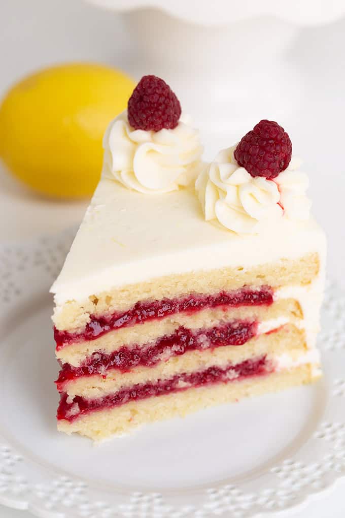 A slice of layered cake with zesty lemon notes, adorned with white frosting and two juicy raspberries on top, resting elegantly on a white plate.