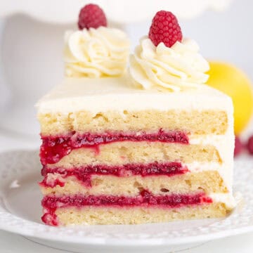 A slice of layered cake with zesty lemon frosting, raspberry filling, and a raspberry topped with cream sits elegantly on a white plate.
