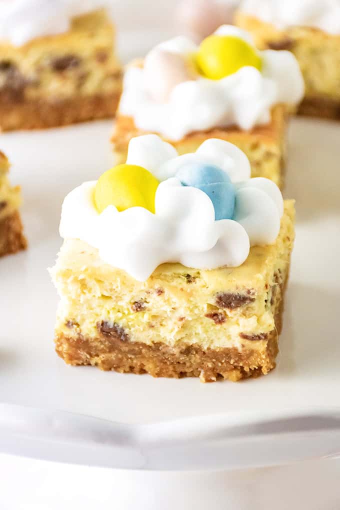 A delectable cheesecake bar topped with white frosting and vibrant yellow and blue Easter eggs sits elegantly on a white surface. This Easter treat boasts a rich brown crust adorned with chocolate chips, making it an irresistible delight for the holiday.
