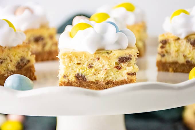 Cheesecake bars artfully topped with whipped cream and colorful candy, reminiscent of festive Easter eggs, presented on a pristine white cake stand.