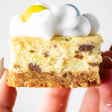 Close-up of a hand holding a slice of Mini Egg Cheesecake with a cookie crust, topped with whipped cream and colorful candies.