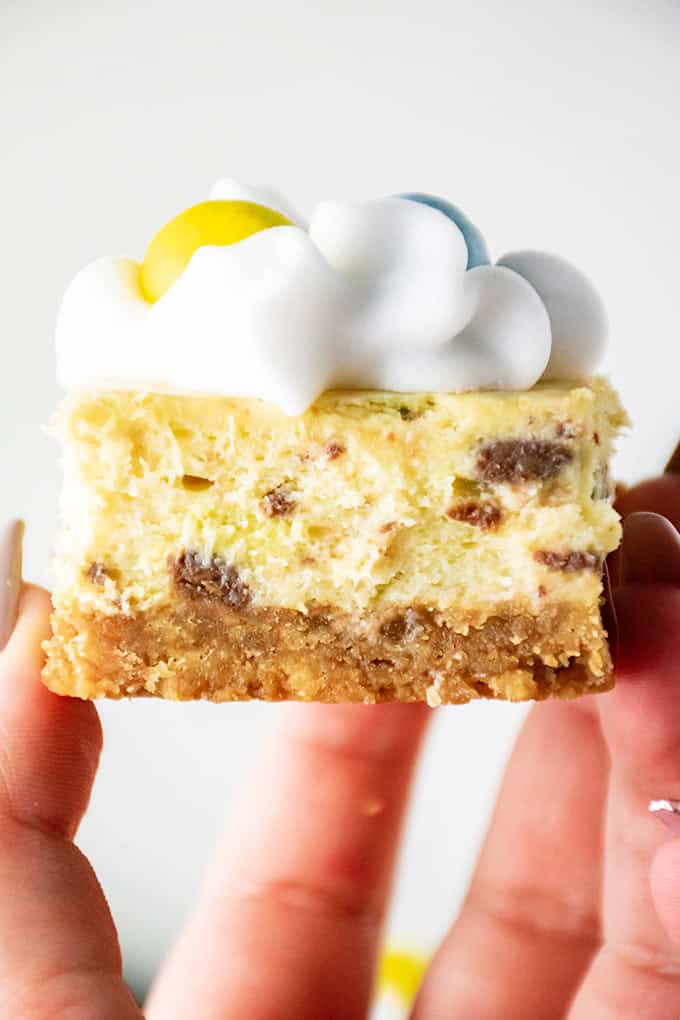 Close-up of a hand holding a slice of Mini Egg Cheesecake with a cookie crust, topped with whipped cream and colorful candies.