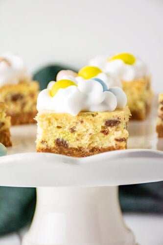Close-up of Mini Egg Cheesecake Bars with a crumbly base, creamy filling speckled with chocolate chips, and a whipped topping adorned with vibrant yellow and blue candy pieces, all elegantly displayed on a white cake stand.